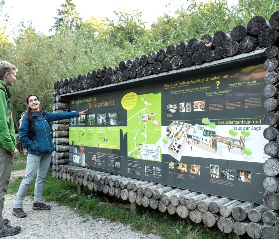 Zwei Personen stehen vor einer Informationstafel im Naturparkzentrum Teufelsschlucht. Sie zeigt Karten und Informationen über die Umgebung. Zwei Personen stehen vor einer Informationstafel im Naturparkzentrum Teufelsschlucht. Sie zeigt Karten und Informationen über die Umgebung.