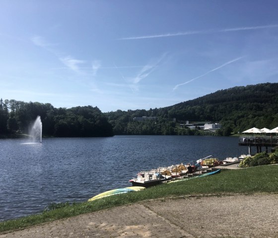 Der Bitburger Stausee in Biersdorf am See mit Tretbooten, einem Springbrunnen und umliegenden Hügeln unter blauem Himmel., © TI Bitburger Land Der Bitburger Stausee in Biersdorf am See mit Tretbooten, einem Springbrunnen und umliegenden Hügeln unter blauem Himmel., © TI Bitburger Land