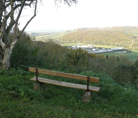 Holzbank auf einer Wiese mit Blick auf eine hügelige Landschaft und ein Industriegebiet in der Ferne. Ein Baum steht links im Bild., © Felsenland Südeifel Tourismus GmbH, Christian Calonec-Rauchfuss Holzbank auf einer Wiese mit Blick auf eine hügelige Landschaft und ein Industriegebiet in der Ferne. Ein Baum steht links im Bild., © Felsenland Südeifel Tourismus GmbH, Christian Calonec-Rauchfuss