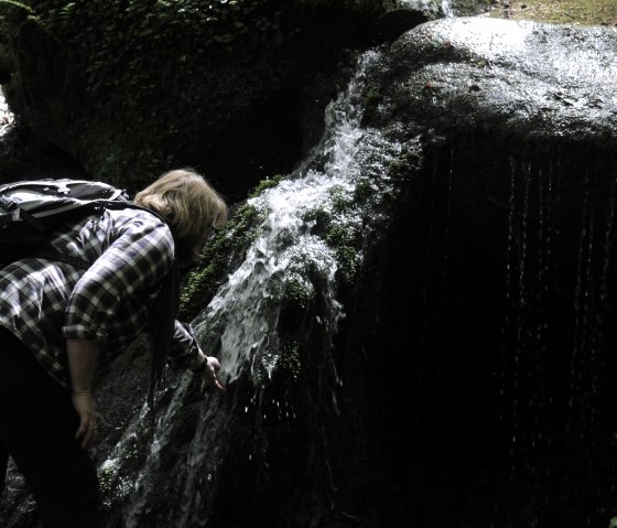 A person with a rucksack bends over a small, moss-covered waterfall in the forest., © Felsenland Südeifel Tourismus GmbH A person with a rucksack bends over a small, moss-covered waterfall in the forest., © Felsenland Südeifel Tourismus GmbH