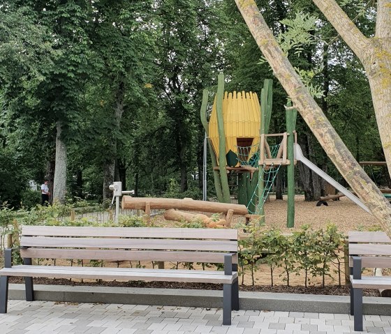 Spielplatz im Park Maximiner Wäldchen in Bitburg mit Bänken, Klettergerüst und Rutsche, umgeben von Bäumen und Grünflächen., © TI Bitburger Land Spielplatz im Park Maximiner Wäldchen in Bitburg mit Bänken, Klettergerüst und Rutsche, umgeben von Bäumen und Grünflächen., © TI Bitburger Land
