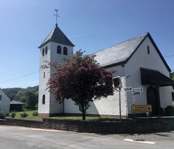 Weiße Kirche mit Turm und rotem Baum im Vordergrund, blauer Himmel. Straßenschilder weisen nach Arzfeld und Koosbüsch., © Fellmann Weiße Kirche mit Turm und rotem Baum im Vordergrund, blauer Himmel. Straßenschilder weisen nach Arzfeld und Koosbüsch., © Fellmann