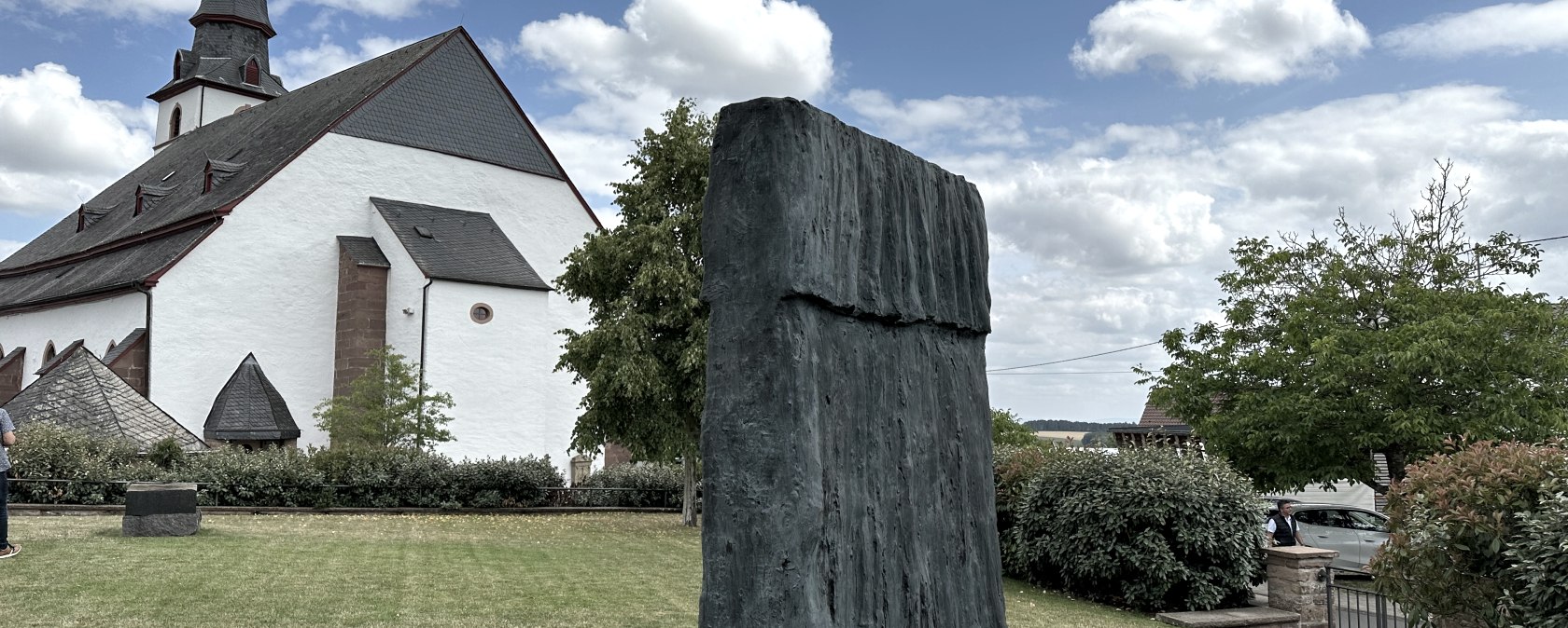 Kirche mit spitzem Turm, moderne Skulptur im Vordergrund, grüner Rasen, Bäume und blauer Himmel mit Wolken. Kirche mit spitzem Turm, moderne Skulptur im Vordergrund, grüner Rasen, Bäume und blauer Himmel mit Wolken.