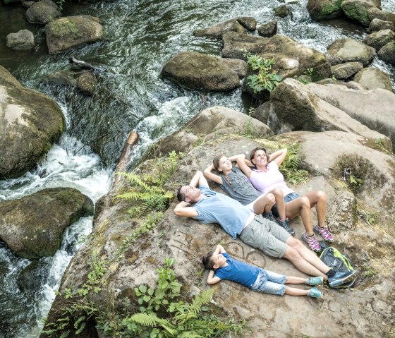 A family lies relaxed on a large rock in the middle of a flowing stream with waterfalls and surrounded by lush greenery., © Felsenland Südeifel Tourismus GmbH A family lies relaxed on a large rock in the middle of a flowing stream with waterfalls and surrounded by lush greenery., © Felsenland Südeifel Tourismus GmbH