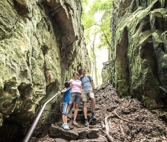 Eine Familie wandert durch die Teufelsschlucht, umgeben von hohen, moosbewachsenen Felswänden und Bäumen. Eine Familie wandert durch die Teufelsschlucht, umgeben von hohen, moosbewachsenen Felswänden und Bäumen.
