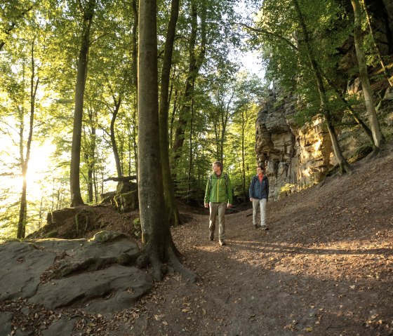 Two hikers are walking on a forest path along a rock face. The sun shines through the trees and bathes the scene in warm light., © Dominik Ketz Two hikers are walking on a forest path along a rock face. The sun shines through the trees and bathes the scene in warm light., © Dominik Ketz