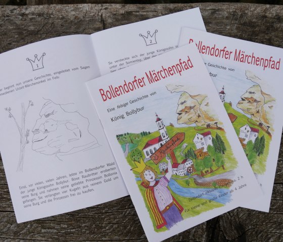 Malbuchseiten des Bollendorfer Märchenpfads mit bunten Illustrationen und Text über König Bollybur auf einem Holztisch., © Felsenland Südeifel Tourismus GmbH Malbuchseiten des Bollendorfer Märchenpfads mit bunten Illustrationen und Text über König Bollybur auf einem Holztisch., © Felsenland Südeifel Tourismus GmbH