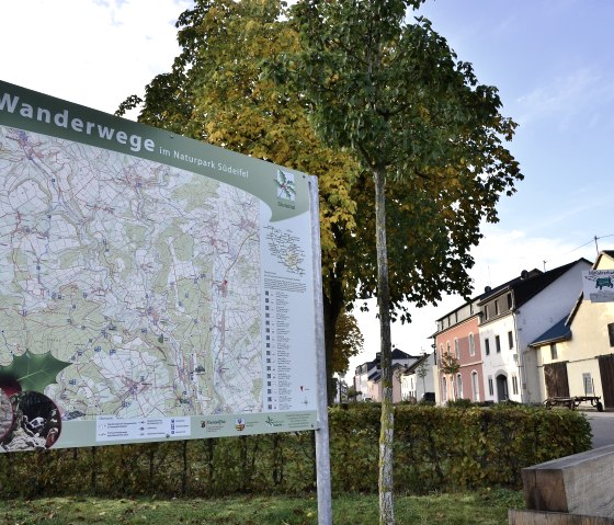 Wanderkarte im Naturpark Südeifel vor herbstlichen Bäumen und Häusern in Wolsfeld. Eine Ruhebank und ein Briefkasten sind ebenfalls zu sehen., © TI Bitburger Land Wanderkarte im Naturpark Südeifel vor herbstlichen Bäumen und Häusern in Wolsfeld. Eine Ruhebank und ein Briefkasten sind ebenfalls zu sehen., © TI Bitburger Land