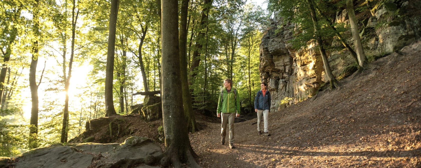 Two hikers are walking on a forest path along a rock face. The sun shines through the trees and bathes the scene in warm light., © Dominik Ketz Two hikers are walking on a forest path along a rock face. The sun shines through the trees and bathes the scene in warm light., © Dominik Ketz