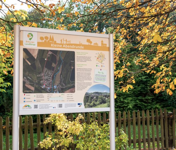 Schild zur Wanderroute 'Kleine Abendrunde' im Bitburger Land, umgeben von herbstlichen Bäumen und einem Holzzaun. Schild zur Wanderroute 'Kleine Abendrunde' im Bitburger Land, umgeben von herbstlichen Bäumen und einem Holzzaun.