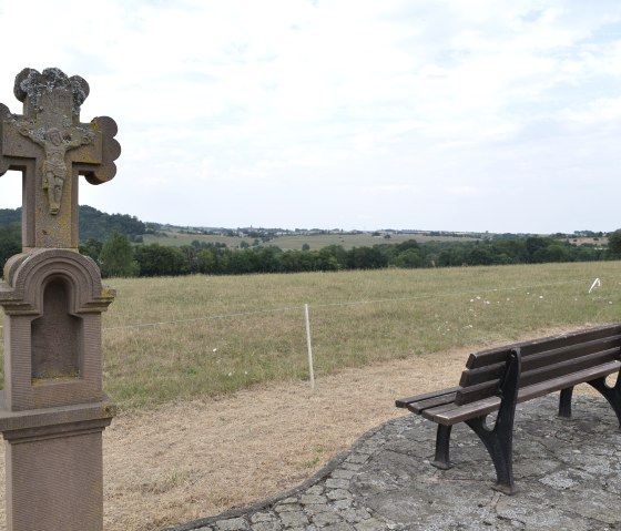 Bank und steinernes Kreuz in ländlicher Landschaft mit weitem Blick über Felder und Hügel., © TI Bitburger Land Bank und steinernes Kreuz in ländlicher Landschaft mit weitem Blick über Felder und Hügel., © TI Bitburger Land