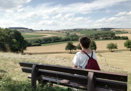 Eine Person mit rotem Rucksack sitzt auf einer Bank und blickt auf eine weite Landschaft mit Feldern und Bäumen unter einem blauen Himmel., © TI Bitburger Land Eine Person mit rotem Rucksack sitzt auf einer Bank und blickt auf eine weite Landschaft mit Feldern und Bäumen unter einem blauen Himmel., © TI Bitburger Land