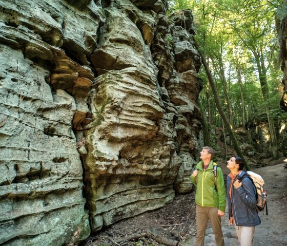 Zwei Wanderer stehen vor beeindruckenden Felsformationen in einem Wald. Die Felsen haben markante, geschichtete Strukturen. Sonnenlicht fällt durch die Bäume. Zwei Wanderer stehen vor beeindruckenden Felsformationen in einem Wald. Die Felsen haben markante, geschichtete Strukturen. Sonnenlicht fällt durch die Bäume.