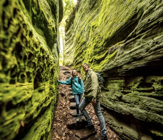 Zwei Wanderer in einer engen, moosbedeckten Schlucht. Die Wände sind grün und steil, der Boden ist mit Laub bedeckt. Die Szene wirkt abenteuerlich., © Eifel Tourismus GmbH, Dominik Ketz Zwei Wanderer in einer engen, moosbedeckten Schlucht. Die Wände sind grün und steil, der Boden ist mit Laub bedeckt. Die Szene wirkt abenteuerlich., © Eifel Tourismus GmbH, Dominik Ketz