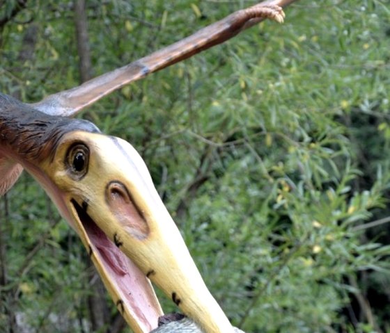 Ein Flugsaurier-Modell hält einen Fisch im Schnabel, umgeben von grüner Vegetation im Dinopark. Ein Flugsaurier-Modell hält einen Fisch im Schnabel, umgeben von grüner Vegetation im Dinopark.