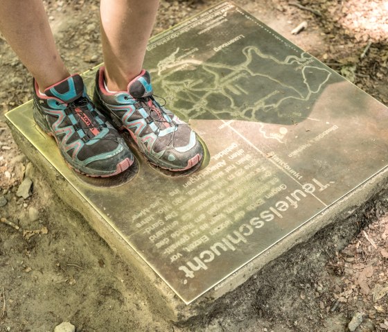 A person with colorful sports shoes stands on an information board in the forest. The board shows a map and the text 'Teufelsschlucht' (Devil's Gorge)., © Felsenland Südeifel Tourismus GmbH A person with colorful sports shoes stands on an information board in the forest. The board shows a map and the text 'Teufelsschlucht' (Devil's Gorge)., © Felsenland Südeifel Tourismus GmbH