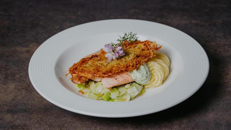 Une belle assiette de saumon grillé, entourée de purée et de poireau. Le plat est décoré d'herbes et d'une fleur comestible.