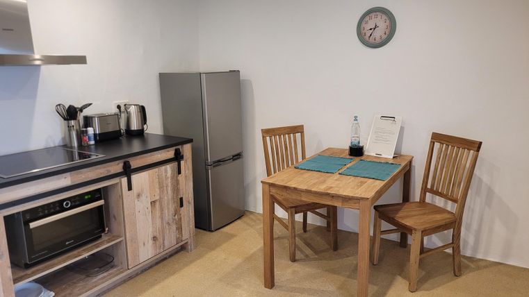 Eine moderne Küche mit einem Esstisch und zwei Stühlen. Im Hintergrund sind ein Kühlschrank und verschiedene Küchengeräte zu sehen.