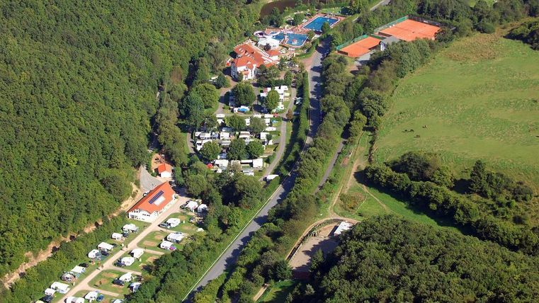 Une vue aérienne d'un camping entouré de forêt. Il y a des caravanes, une piscine et des courts de tennis à proximité.