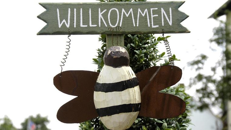 Een bord met de tekst "WELKOM" en een houten figuur van een bij. De bij heeft zwarte en witte strepen en naar buiten gerichte vleugels.