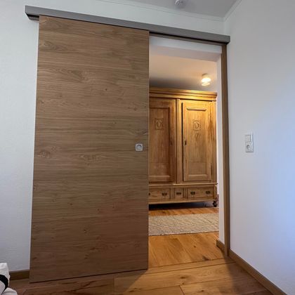 Eine Holzschiebetür öffnet sich in einen Raum mit Holzschränken. Der Boden ist aus Holz und der Raum wirkt einladend.