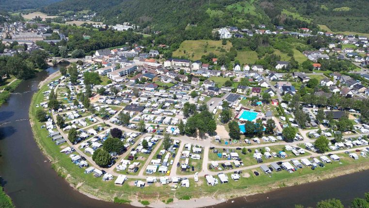 Une vue aérienne d'un camping au bord d'une rivière. Entouré de collines verdoyantes et de maisons, le site est agrémenté d'arbres et d'une piscine.