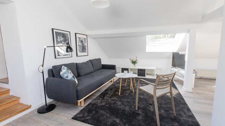 Ein modernes Wohn- und Esszimmer mit einem grauen Sofa und einem hellen Tisch. Die Wände sind weiß und mit Bildern dekoriert, der Boden ist aus Holz.