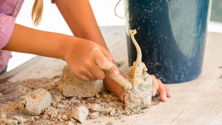Een kind vormt zand met één hand terwijl het een kleine figuur vasthoudt. Op de achtergrond staat een blauwe emmer.