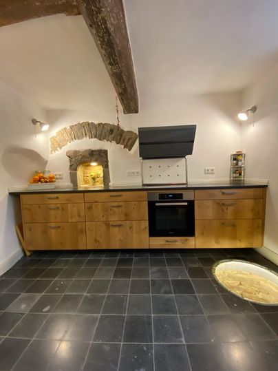 Een moderne keuken met houten kasten en een zwart fornuis. Op de achtergrond is een rustieke steenoven zichtbaar.