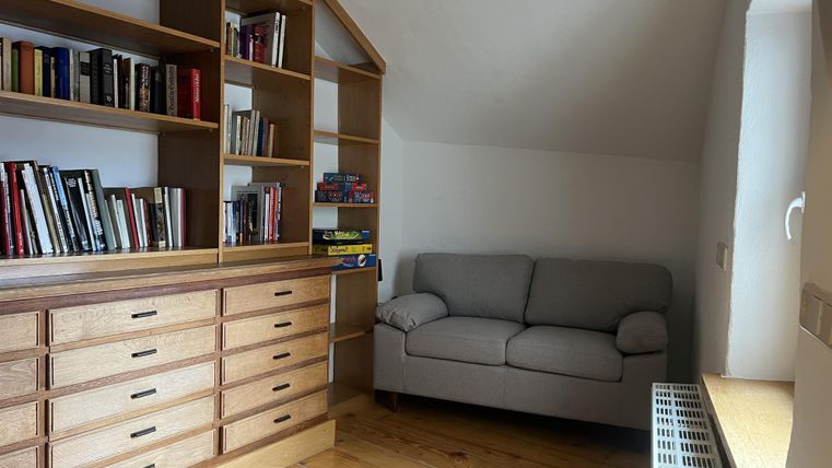 Een gezellige kamer met een boekenplank en een grijze bank. De vloer is van hout, en het licht valt zachtjes door het raam.