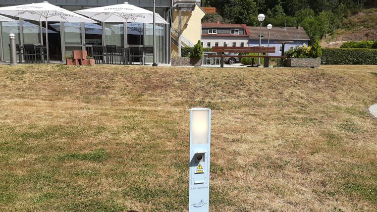 A bright, modern light fixture stands on a lawn in front of a building. In the background, tablecloths and umbrellas of an outdoor area are visible.