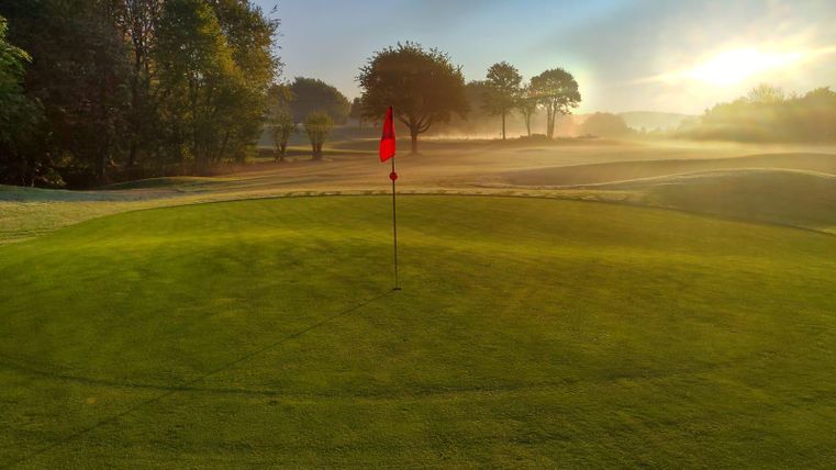 Un parcours de golf au soleil du matin avec un mât de drapeau au centre. Le brouillard confère à la scène une atmosphère calme et paisible.