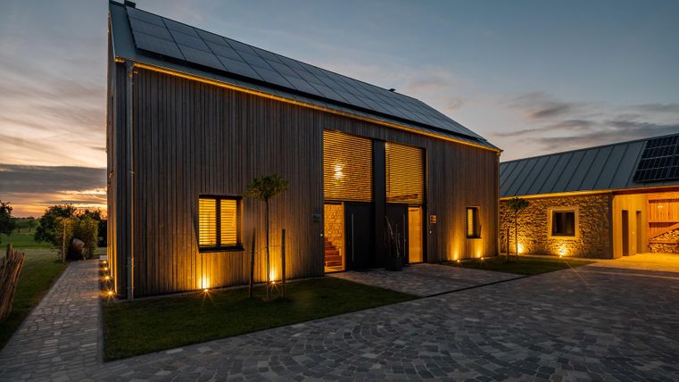 Ein modernes Haus mit Holzverkleidung und Solarpanelen, beleuchtet bei Sonnenuntergang. Die Fassade hat große Fenster und ist von einer Einfahrt mit Steinen umgeben.