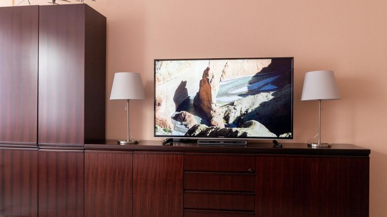 Ein modernes Wohnzimmer mit einem TV auf einem Holzschrank. Zwei Tischlampen stehen neben dem Fernseher und die Wände sind in einem sanften Farbton gestrichen.