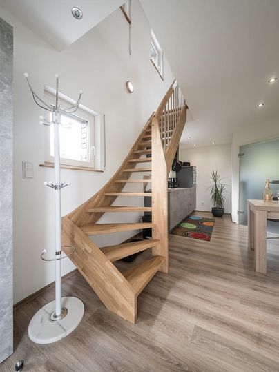 Une cage d'escalier moderne avec un escalier en bois et beaucoup de lumière naturelle. Au premier plan, il y a un porte-manteau et à l'arrière-plan, on voit une cuisine ouverte.