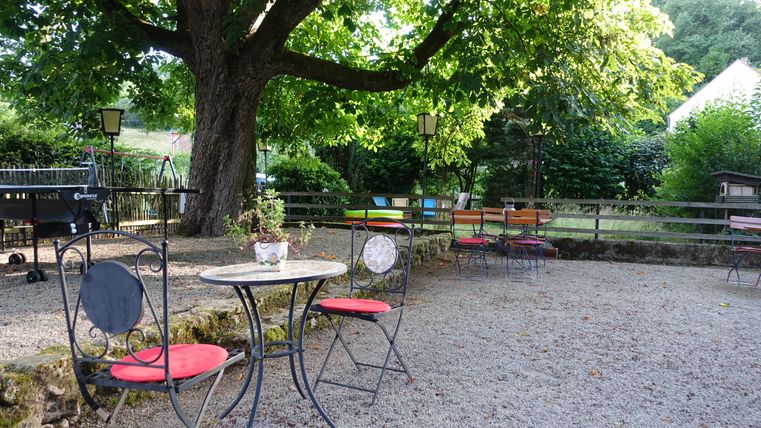 Un espace extérieur confortable avec une table ronde et des chaises sous un grand arbre. Plusieurs chaises colorées sont disposées autour de l'espace, entourées de feuillage vert.