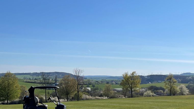 Ein Golfwagen steht auf einem grünen Feld mit einer weitläufigen Landschaft im Hintergrund. Der Himmel ist klar und blau, und die Bäume sprießen im Frühling.