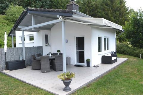 Ein modernes, kleines Haus mit einer überdachten Terrasse. Es ist von einem gepflegten Garten und Bäumen umgeben.