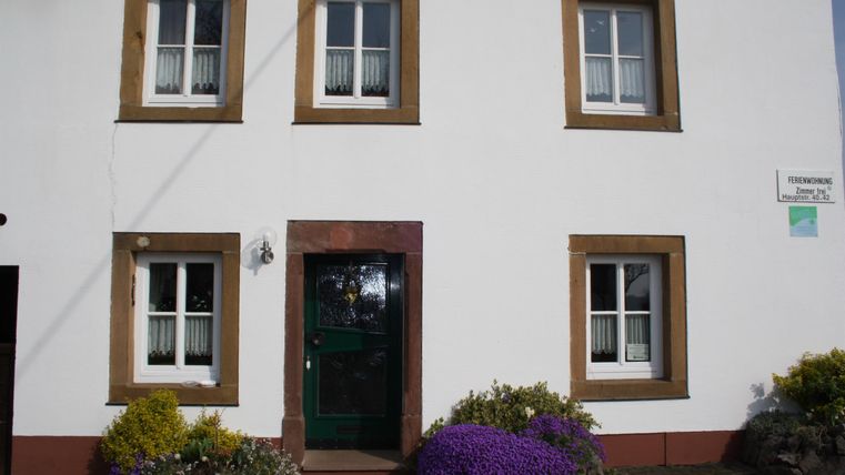 A charming two-story house with white walls and brown window frames. Colorful plants bloom in front of the entrance.