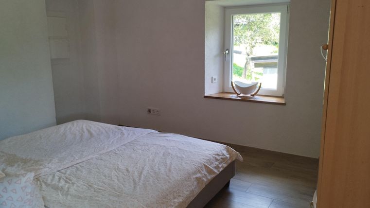 Een eenvoudige slaapkamer met een groot bed en helder daglicht dat door een raam stroomt. De wanden zijn wit en de vloer is van hout.