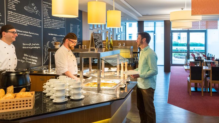 A modern restaurant area with a buffet. A guest is standing at the buffet and is being served by two chefs.