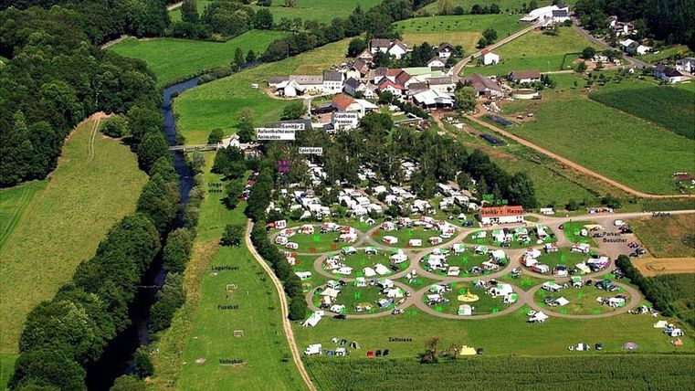 Ein Luftbild eines Campingplatzes mit vielen Zelten und Wohnmobilen in einer grünen Landschaft. Im Hintergrund fließt ein kleiner Fluss und es sind einige Gebäude zu sehen.