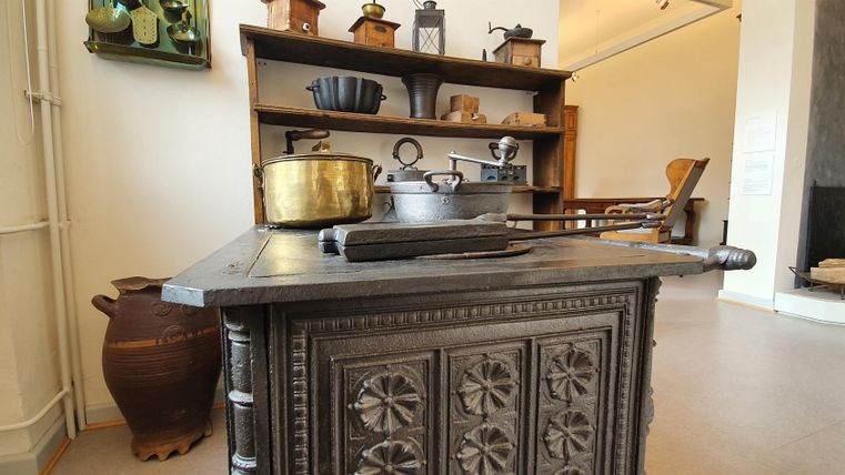 A historical table with intricate decorations stands in the foreground. In the background, various kitchen utensils and a shelf with additional objects can be seen.