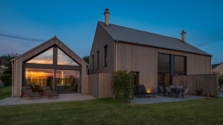 Ein modernes Holzhaus mit großen Fenstern, das zu einem stilvollen Außenbereich führt. Der Himmel ist in der Dämmerung beleuchtet, was eine gemütliche Atmosphäre schafft.