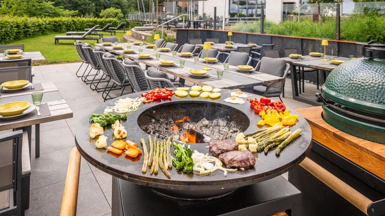 Une station de grill moderne en plein air, entourée d'un jardin bien entretenu. Sur la table, une variété de légumes et de viandes sont préparés pour le barbecue.