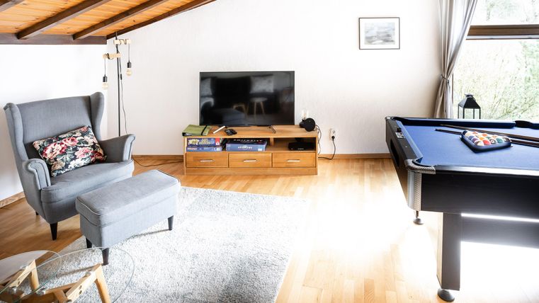 A modern living room with a cozy armchair and a billiard table. The room has large windows and a warm wooden ceiling.