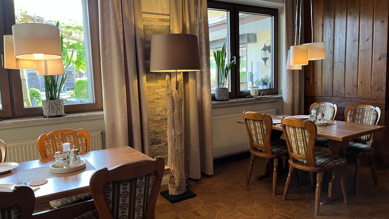 Ein gemütlicher Innenraum mit Holztischen und Stühlen. Große Fenster sorgen für viel Tageslicht und eine angenehme Atmosphäre.