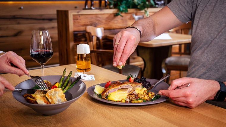 Zwei Teller mit verschiedenen Gerichten auf einem Tisch. Ein Glas Rotwein und ein Bier stehen daneben.