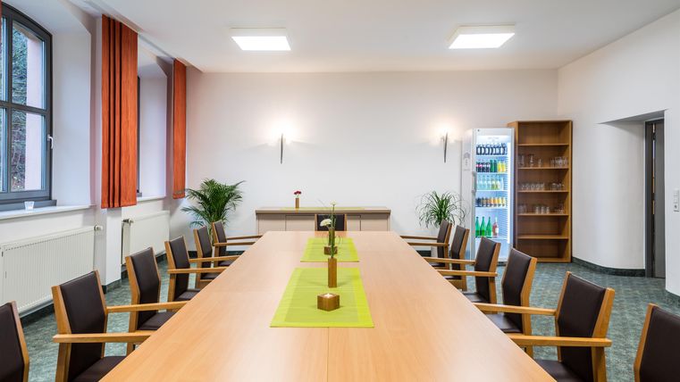 Een moderne vergaderruimte met een lange tafel en stoelen. De muren zijn licht en er zijn planten en boekenplanken op de achtergrond.