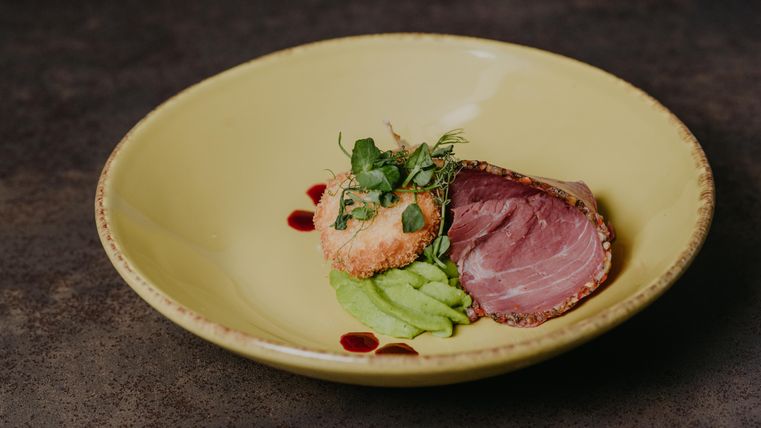 Un plat élégant sur une assiette jaune, composé de viande tendre, d'une croûte croustillante et d'une purée verte. Décoré avec des herbes fraîches et une sauce rouge.